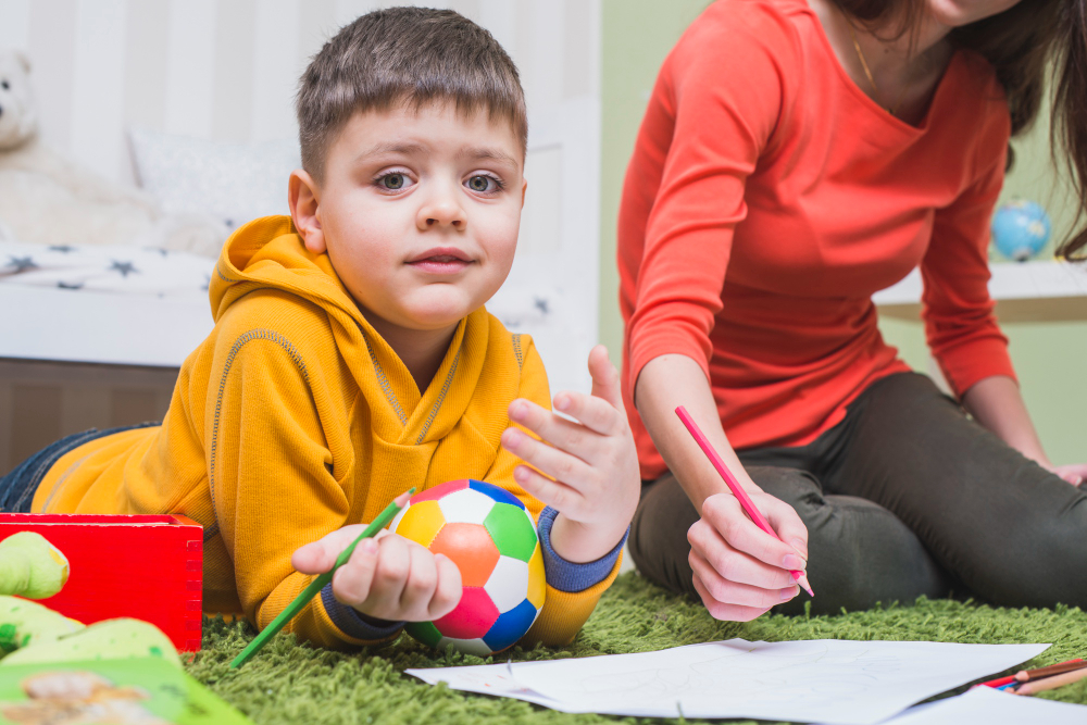 A young boy in a hoodie lying on a rug, holding a colorful ball, with a woman in an orange shirt next to him drawing on a piece of paper. The scene suggests learning activities in a cozy indoor environment, and illustrate the promotion of the non-profit association "Surfing Autism" in Dominican Republic